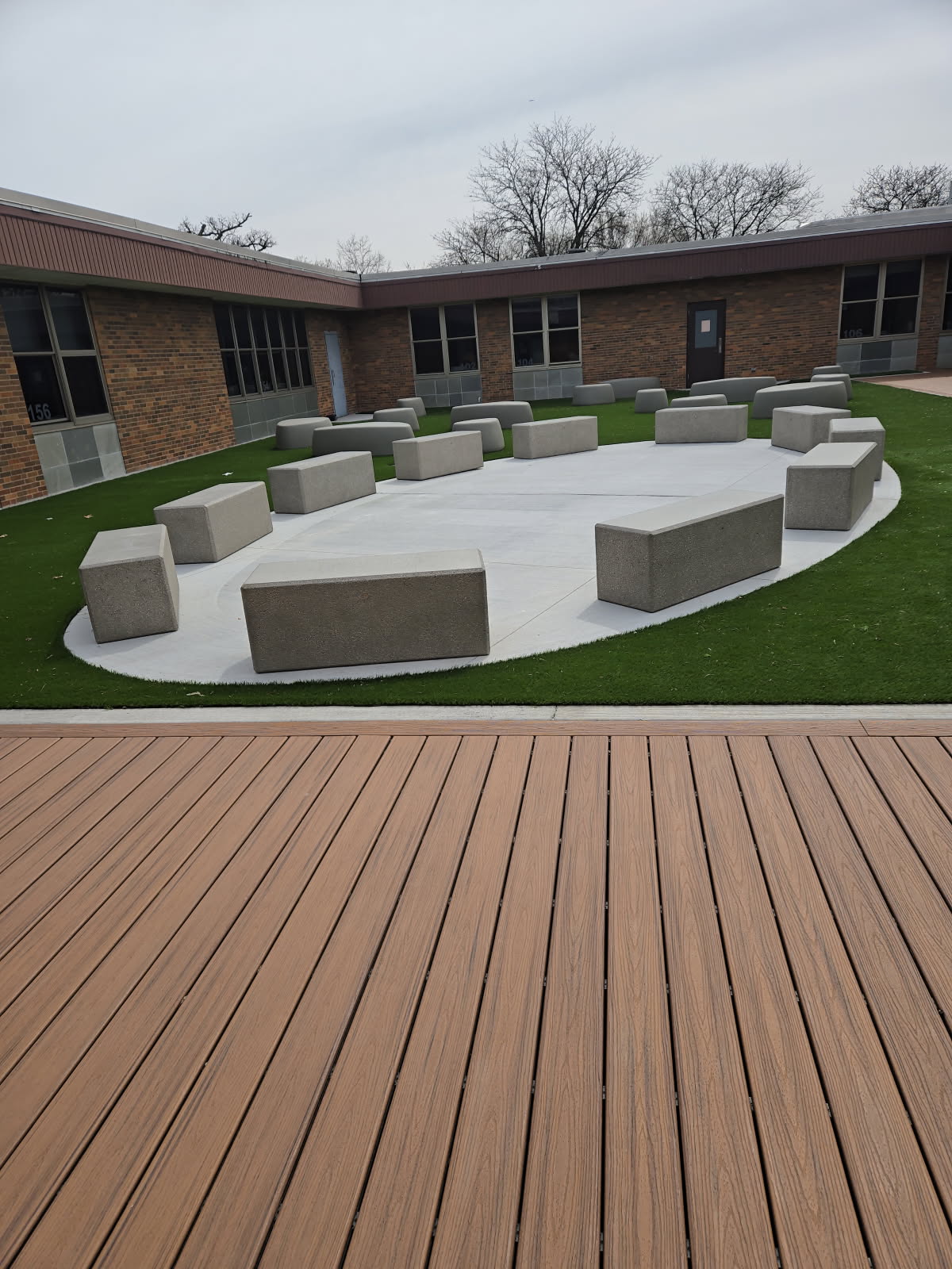 Modern concrete benches arranged in a circular outdoor seating area at Bensenville Blackhawk School courtyard, featuring artificial turf and composite wood decking.