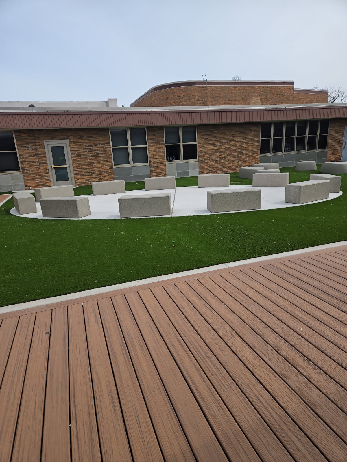 Modern outdoor seating area at Bensenville Blackhawk School featuring concrete benches arranged on a circular concrete pad surrounded by artificial turf and composite wood decking