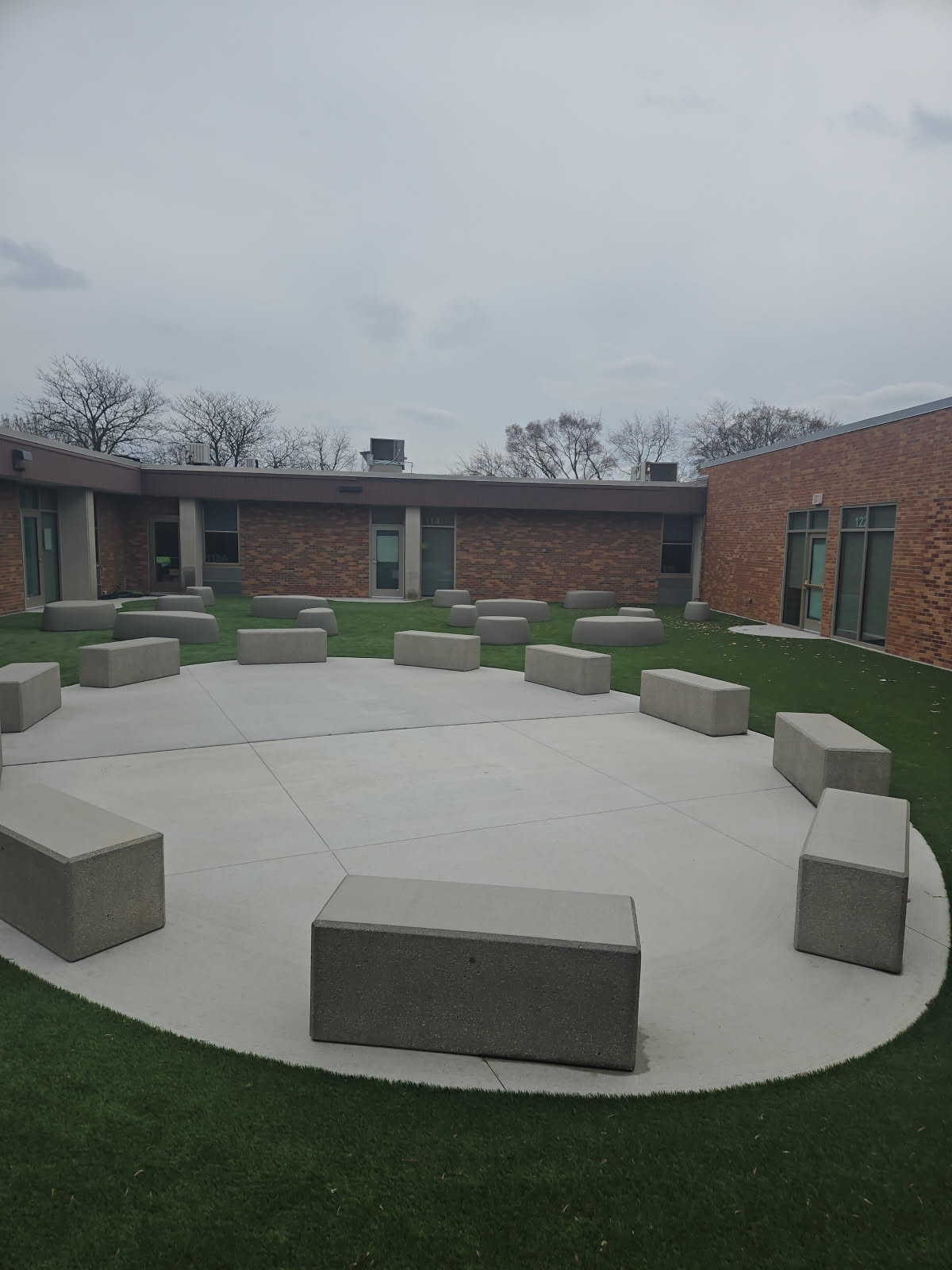 Outdoor circular seating area at Bensenville Blackhawk School with evenly spaced concrete benches on a concrete pad surrounded by artificial turf, designed for collaborative learning and gatherings.