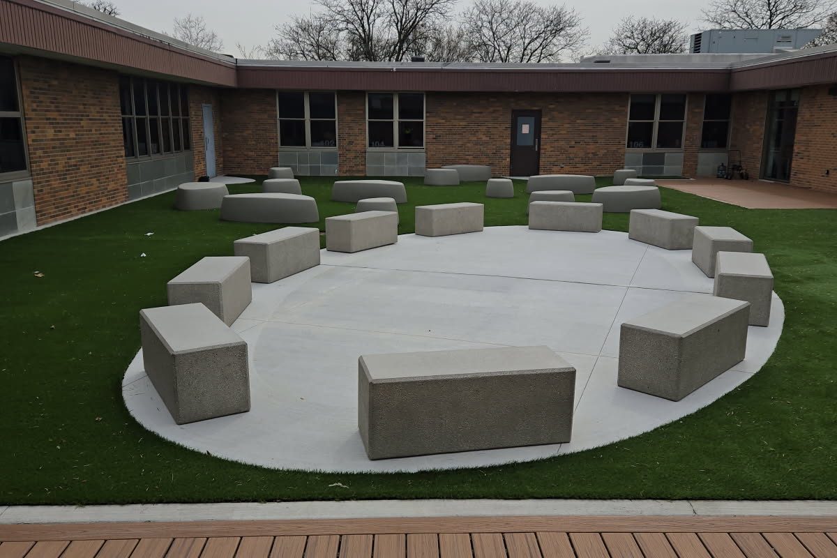 Modern circular concrete bench seating area at Bensenville Blackhawk School, surrounded by artificial turf and wooden decking with brick school buildings in the background.